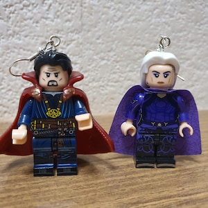 May include: Two Lego-style earrings featuring superhero characters. One has a red cape and black hair, the other has a purple cape and white hair. Both have silver earring hooks.