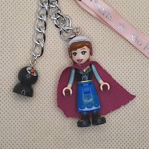 Puede incluir: Una cadena plateada con dos colgantes. Uno es una figura negra con un dise&ntilde;o naranja y blanco. El otro es una figura estilo Lego de un personaje que lleva una capa morada, una falda azul y un sombrero blanco. Una cinta rosa est&aacute; en el fondo.