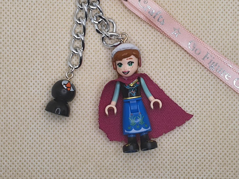 Puede incluir: Una cadena plateada con dos colgantes. Uno es una figura negra con un dise&ntilde;o naranja y blanco. El otro es una figura estilo Lego de un personaje que lleva una capa morada, una falda azul y un sombrero blanco. Una cinta rosa est&aacute; en el fondo.