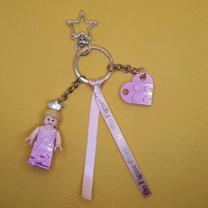 Può includere: Un portachiavi argentato con un ciondolo a forma di stella, un ciondolo a forma di cuore rosa e una figura di principessa Lego. Il portachiavi presenta anche un nastro rosa con la scritta "Go Figure Crafts".