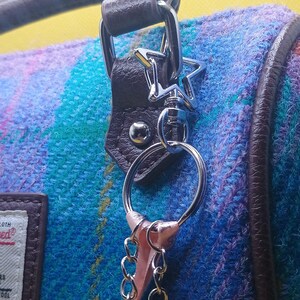 Puede incluir: Primer plano de un bolso con un colorido estampado de cuadros en tonos azules, morados y verdes. El bolso presenta un asa y ribetes de cuero marr&oacute;n, con un llavero plateado en forma de estrella.