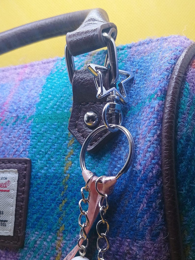 Puede incluir: Primer plano de un bolso con un colorido estampado de cuadros en tonos azules, morados y verdes. El bolso presenta un asa y ribetes de cuero marr&oacute;n, con un llavero plateado en forma de estrella.