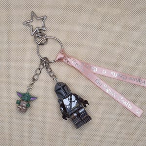 Puede incluir: Un llavero plateado con una parte superior en forma de estrella, que contiene dos colgantes de personajes y una cinta rosa. Un colgante es una figura verde y marr&oacute;n, el otro es una figura gris y negra. La cinta tiene el texto "Go Figure Crafts".