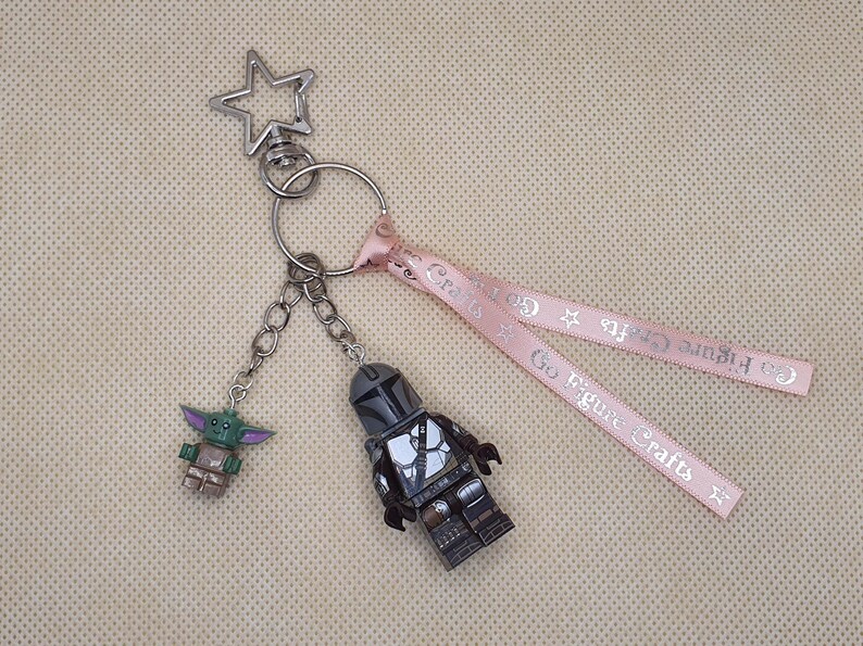 Puede incluir: Un llavero plateado con una parte superior en forma de estrella, que contiene dos colgantes de personajes y una cinta rosa. Un colgante es una figura verde y marr&oacute;n, el otro es una figura gris y negra. La cinta tiene el texto "Go Figure Crafts".