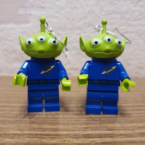 May include: A pair of green alien Lego minifigure earrings. The aliens have three eyes and are wearing blue jumpsuits with a planet logo on the chest.