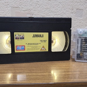 Puede incluir: Una cinta VHS negra de la película "Jumanji" con una etiqueta amarilla. La etiqueta muestra el título de la película y detalles de producción. Junto a ella, un controlador de luz transparente a pilas con varias configuraciones.