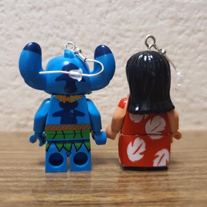 Może przedstawiać: Dwie małe, kolorowe figurki Sticha i Lilo z filmu animowanego Lilo & Stitch. Stitch jest niebieski z dużymi uszami i ż&oacute;łtym naszyjnikiem. Lilo ma na sobie czerwoną sukienkę z białymi kwiatowymi wzorami. Oba mają srebrne pętle do zawieszania.