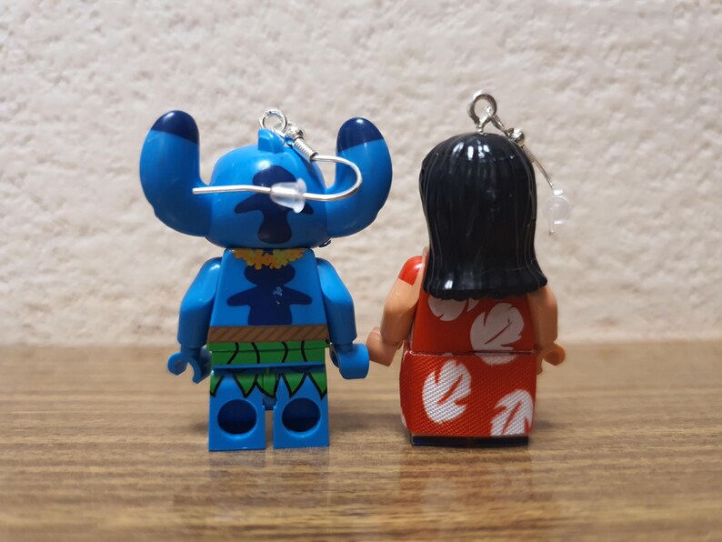 Może przedstawiać: Dwie małe, kolorowe figurki Sticha i Lilo z filmu animowanego Lilo & Stitch. Stitch jest niebieski z dużymi uszami i ż&oacute;łtym naszyjnikiem. Lilo ma na sobie czerwoną sukienkę z białymi kwiatowymi wzorami. Oba mają srebrne pętle do zawieszania.