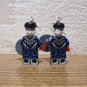 May include: A pair of Lego figure earrings. The figures are dressed in blue and white uniforms with black helmets and silver earring hooks. The earrings are displayed on a wooden surface. The figures are approximately 4 cm tall.
