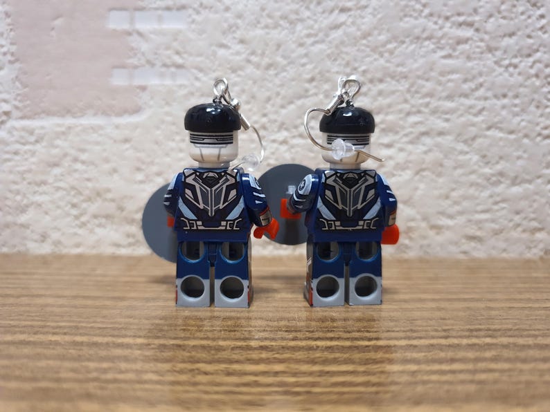 May include: A pair of Lego figure earrings. The figures are dressed in blue and white uniforms with black helmets and silver earring hooks. The earrings are displayed on a wooden surface. The figures are approximately 4 cm tall.