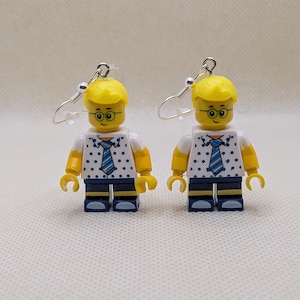 Può includere: Un paio di orecchini pendenti con figure giocattolo in plastica gialla con capelli biondi, occhiali verdi e camicie bianche a pois blu con cravatte a righe blu. Le figure hanno braccia gialle e pantaloni blu scuro, fissate a ganci in metallo argentato.