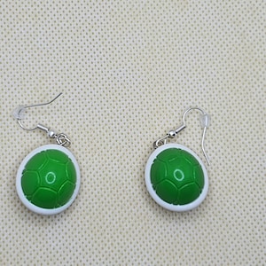 Puede incluir: Un par de pendientes con un diseño de caparazón de tortuga verde con un borde blanco. Los pendientes tienen cierres de gancho plateados. El diseño del caparazón de tortuga es de color verde brillante.