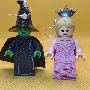 May include: A pair of handmade earrings featuring Lego figures. One earring depicts a green-skinned witch with a black hat and cape. The other earring shows a princess with a silver crown and pink dress. Both earrings have silver hooks.