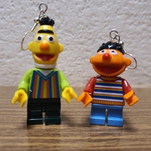 Puede incluir: Un par de pendientes con personajes de Plaza Sésamo. Los pendientes están hechos de plástico y presentan a Bert y Ernie. Bert lleva una camisa a rayas verde y amarilla y Ernie lleva una camisa a rayas roja y azul.