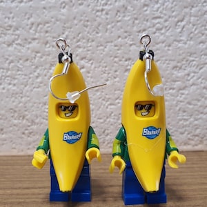 Op de afbeelding: Een paar novelty oorbellen met gele banaanfiguren met zonnebrillen en het woord "Banana!" erop gedrukt. De figuren hebben een blauwe broek en groen-gele mouwen, met zilveren oorhaakjes.