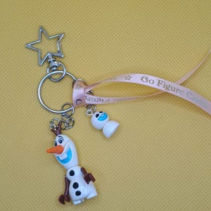 Puede incluir: Un llavero con dos figuras de Olaf de la pel&iacute;cula Frozen. El Olaf m&aacute;s grande tiene una nariz de zanahoria, brazos marrones y botones negros. Un cierre en forma de estrella y una cinta rosa con el texto "Go Figure Craft".