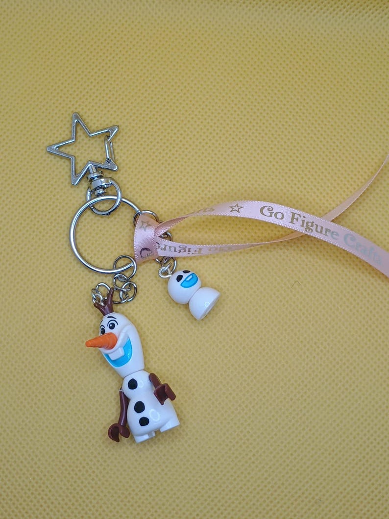 Puede incluir: Un llavero con dos figuras de Olaf de la pel&iacute;cula Frozen. El Olaf m&aacute;s grande tiene una nariz de zanahoria, brazos marrones y botones negros. Un cierre en forma de estrella y una cinta rosa con el texto "Go Figure Craft".
