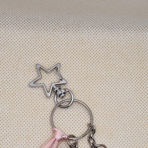 May include: Silver-toned keychain with a star-shaped charm. The keychain includes a split ring, a small pink ribbon, and a chain. The star charm is a five-pointed star with a hollow center. The background is a light beige fabric.