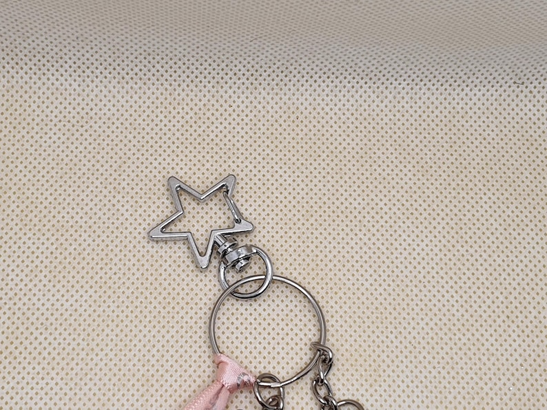 May include: Silver-toned keychain with a star-shaped charm. The keychain includes a split ring, a small pink ribbon, and a chain. The star charm is a five-pointed star with a hollow center. The background is a light beige fabric.