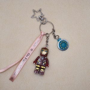 May include: A keychain featuring an Iron Man figure, a circular charm, and a star-shaped clasp. A pink ribbon with the text "Go Figure Crafts" is attached. The Iron Man figure is red, gold, and brown.