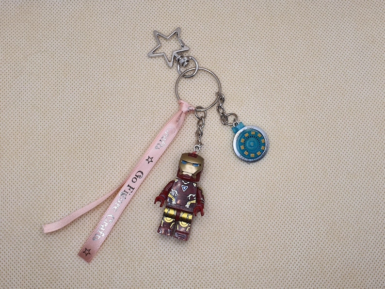 May include: A keychain featuring an Iron Man figure, a circular charm, and a star-shaped clasp. A pink ribbon with the text "Go Figure Crafts" is attached. The Iron Man figure is red, gold, and brown.