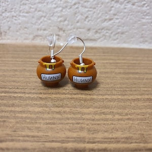 Puede incluir: Un par de pendientes de tarro de miel con la palabra "Hunny" impresa en la parte delantera. Los pendientes están hechos de plástico marrón y tienen ganchos plateados.