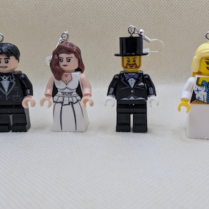 May include: Four dangling earrings featuring plastic toy figures. Two figures wear black formal suits, one wears a white gown with black accents, and one wears a white gown with a blue bodice pattern. Each figure has a silver-toned metal earring hook.