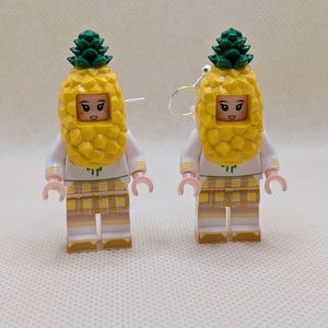 May include: A pair of earrings featuring minifigure characters wearing yellow pineapple costumes with green tops. The figures have white shirts with green details and yellow plaid skirts. Each earring includes a silver-toned metal hook for attachment.