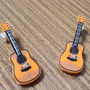 May include: Two miniature acoustic guitars with a distressed orange finish and black strings. The guitars have a small blue and red detail on the body.