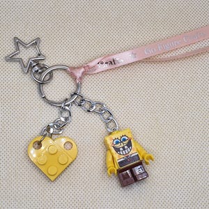 May include: A keychain with a silver star charm, a yellow heart-shaped charm, and a yellow SpongeBob SquarePants figure. The keychain has a pink ribbon with the text "Go Figure Crafts".