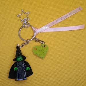 Puede incluir: Un llavero con una figura de bruja estilo Lego con sombrero y capa negros, un dije en forma de corazón verde y un cierre en forma de estrella. Incluye una cinta rosa con el texto "Go Figure Crafts".