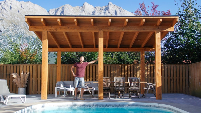May include: A wooden pergola with a flat roof, built over a swimming pool. The pergola is made of natural wood and has a light brown stain. There is a table and chairs under the pergola, and a person is standing near the pool.
