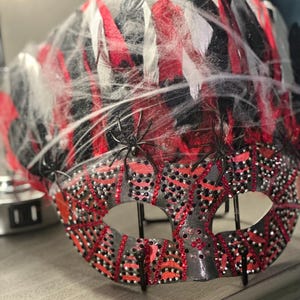 May include: A decorative Halloween mask with a gray base, adorned with red and black feathers, and embellished with red and black rhinestones. The mask is covered with white spiderwebs and features small black spider decorations.