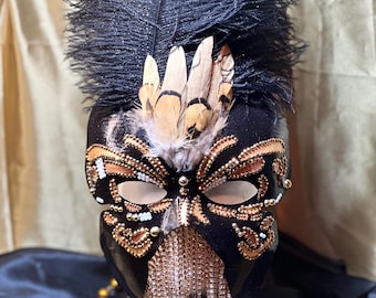 Puede incluir: Una máscara negra de carnaval con adornos dorados y bronce, pedrería y plumas. La máscara presenta un gran penacho de plumas negras brillantes y una cascada de pedrería colgante. Las aberturas para los ojos son ovaladas.