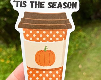 Op de afbeelding: Een sticker met een koffiebekerontwerp met een bruin deksel en oranje stippen. Een pompoenafbeelding staat in het midden van de beker, met de tekst "'TIS THE SEASON" bovenaan. De sticker wordt tegen een vage groene achtergrond gehouden.