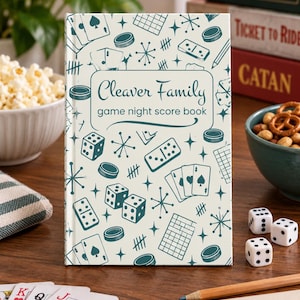 Puede incluir: Un libro de puntuaciones para noches de juegos de tapa dura en color crema, titulado Cleaver Family game night score book en texto verde azulado oscuro, con un patrón repetitivo de cartas, dados, dominós y estrellas.