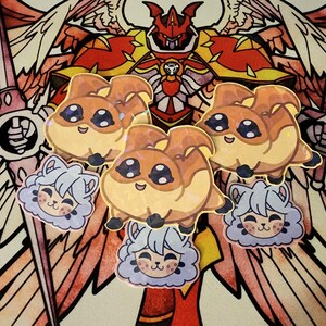 May include: A collection of cartoon stickers featuring a brown and tan creature with large eyes and a smiling face, along with stickers of a light blue cat-like character. The stickers are set against a colorful background.