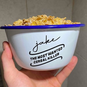 Könnte beinhalten: Weiße Emaille-Schüssel mit blauem Rand, gefüllt mit Müsli. Die Schüssel trägt den Text "jake THE MOST WANTED CEREAL KILLER".