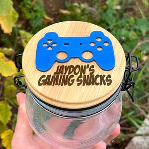 May include: A clear glass jar with a wooden lid featuring a blue game controller design. The lid is engraved with "JAYDON'S GAMING SNACKS". The jar has a metal clasp and is designed for storing snacks. The jar is perfect for storing snacks.