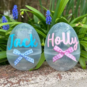 May include: Two clear plastic Easter eggs, each with a name in script: "Jack" in blue and "Holly" in pink. Each egg has a matching bow at the bottom. The background features green foliage and blue flowers.