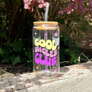 May include: A clear glass can-shaped tumbler with a bamboo lid and a clear straw. The tumbler features the text "COOL UNCLES CLUB" in yellow and purple. Decorative stars and flowers are also present.