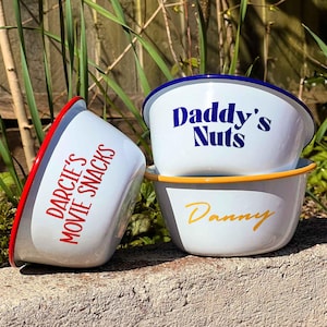May include: Three white enamel bowls with colorful rims. The bowls are stacked on top of each other. The top bowl has blue and white trim and says "Daddy's Nuts" in blue letters. The middle bowl has a yellow rim and says "Danny" in yellow letters. The bottom bowl has a red rim and says "Darcie's Movie Snacks" in red letters.