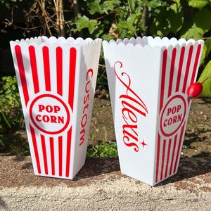 May include: Two white popcorn containers with red stripes and the words "Pop Corn" in a circle. One container has the word "Custom" in red, and the other has the name "Alexis" in red.