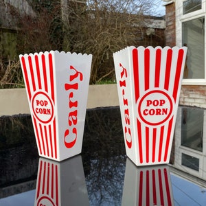 Peut inclure: Deux contenants à popcorn en carton blanc avec des rayures rouges et blanches. Les contenants portent l'inscription "Pop Corn". Les contenants sont posés sur une surface noire.