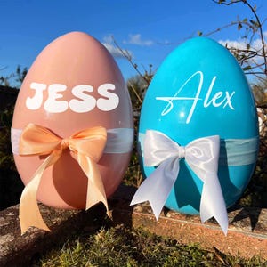 May include: Two large, egg-shaped containers, one peach and one turquoise, are displayed outdoors. The peach egg has the name "JESS" in white, while the turquoise egg has "Alex." Both eggs are adorned with ribbon bows and white ribbon bands.