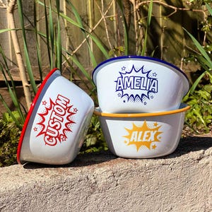 May include: White enamel bowls with coloured rims and custom text. One bowl has a red rim and the word "CUSTOM" in red. Two bowls have names in blue and yellow, with blue and yellow rims. The bowls are stacked on a stone surface.