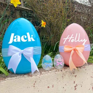 Puede incluir: Dos huevos de Pascua grandes y decorados, uno azul con el nombre "Jack" y otro rosa con el nombre "Holly". Dos huevos más pequeños, uno azul con "Jack" y otro rosa con "Holly", están delante de los huevos más grandes.