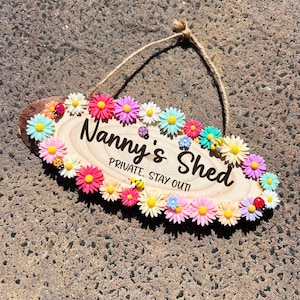 Könnte beinhalten: Ein Holzschild mit dem Text "Nanny's Shed Private Stay Out!" umgeben von bunten Blumen und kleinen dekorativen Insekten.