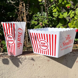 May include: Two white popcorn containers with red stripes and text. One container has "Charlie" in red script. The other has "The Smith's movie night snacks" in red script. Both have a "Pop Corn" logo.
