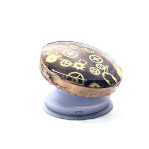 Op de afbeelding: Een bruine en gouden telefoonhouder met een uitklapbare standaard. De houder is versierd met een patroon van gouden tandwielen op een bruine achtergrond.